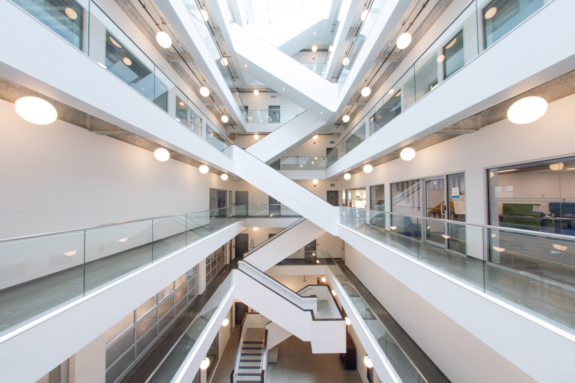 Image of a few of the floors of the Kelowna Innovation Centre, connected by stairs in the background. Offices, boardrooms, and social spaces can be seen on the different floors. Glass and metal railings can be seen on the sides of each floor.